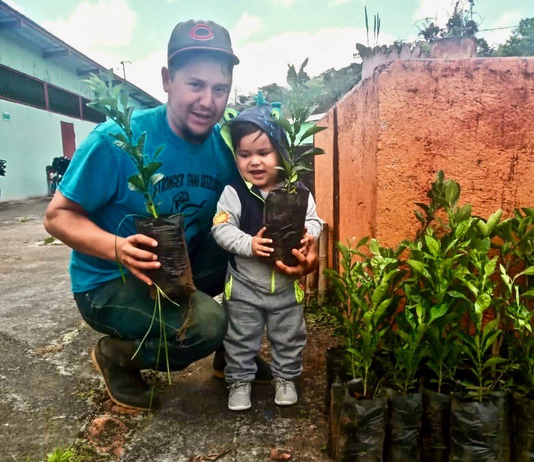 Carlos Evert of SOPPEXCCA cooperative kneels down next to a toddler. They both hold a fruit tree sapling in their hands that they received at a recent tree delivery from Grow Ahead.